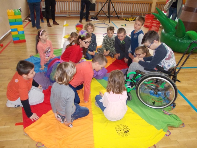 Na zdjęciu jest grupa dzieci, które siedzą na podłodze na kolorowej chuście