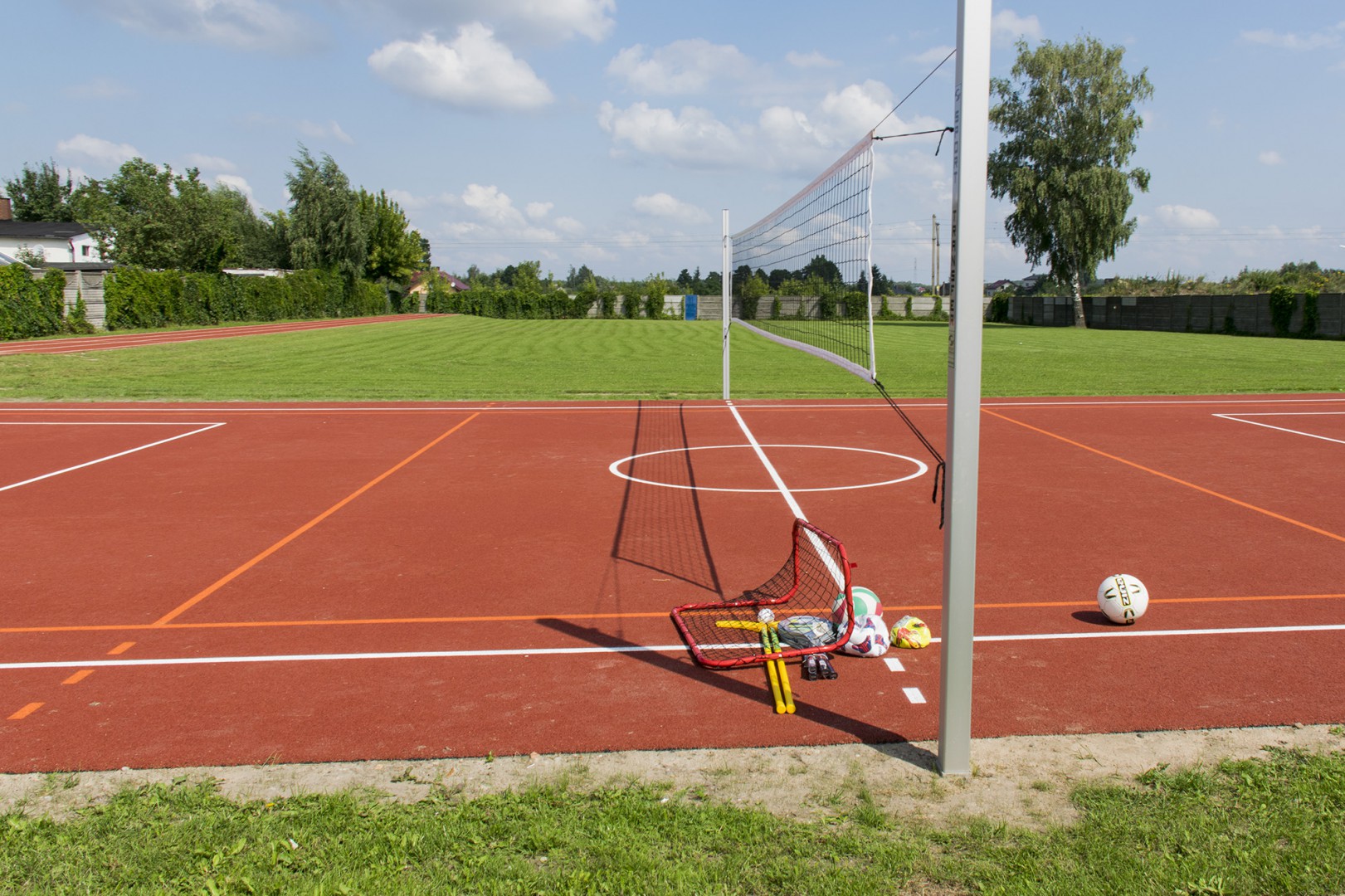 Na zdjęciu widać boisko z czerwoną nawierzchnią, siatkę do siatkówki oraz piłki i małe bramki