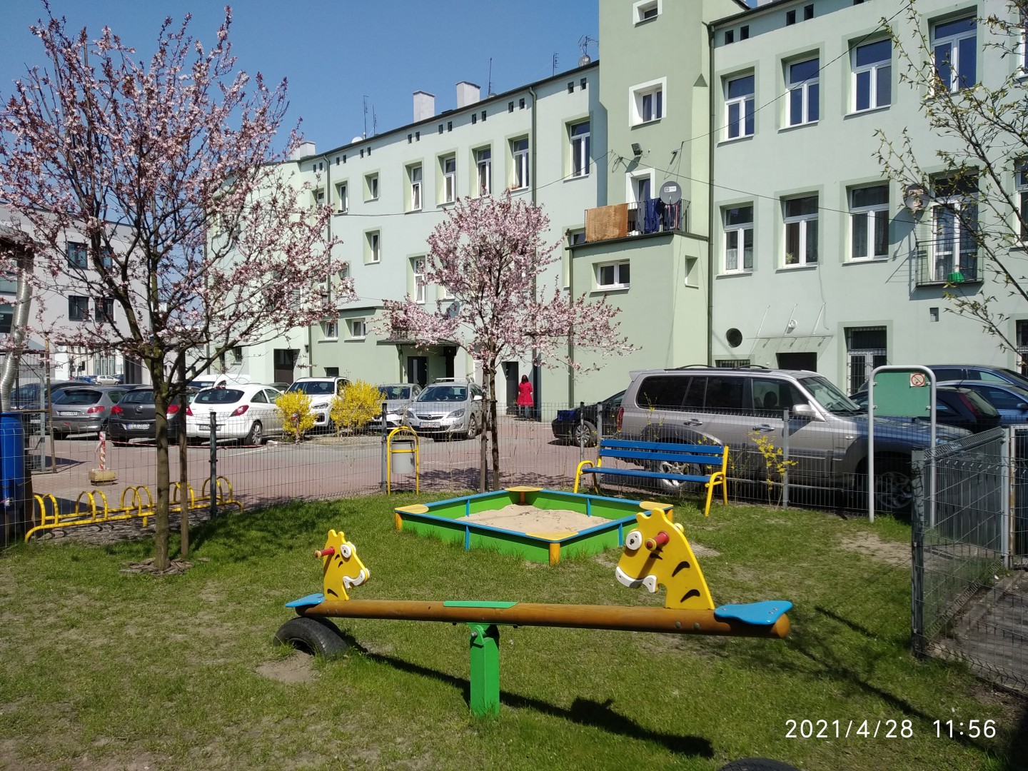 Na zdjęciu widać plac zabaw, ogrodzenie, a za nimi zieloną wysoką kamienicę