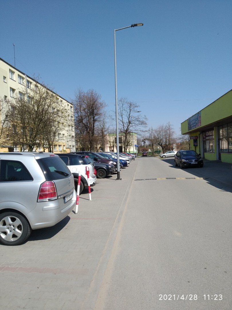 Na zdjęciu widać małą osiedlową uliczkę, parkujące samochody oraz bloki i lampy uliczne