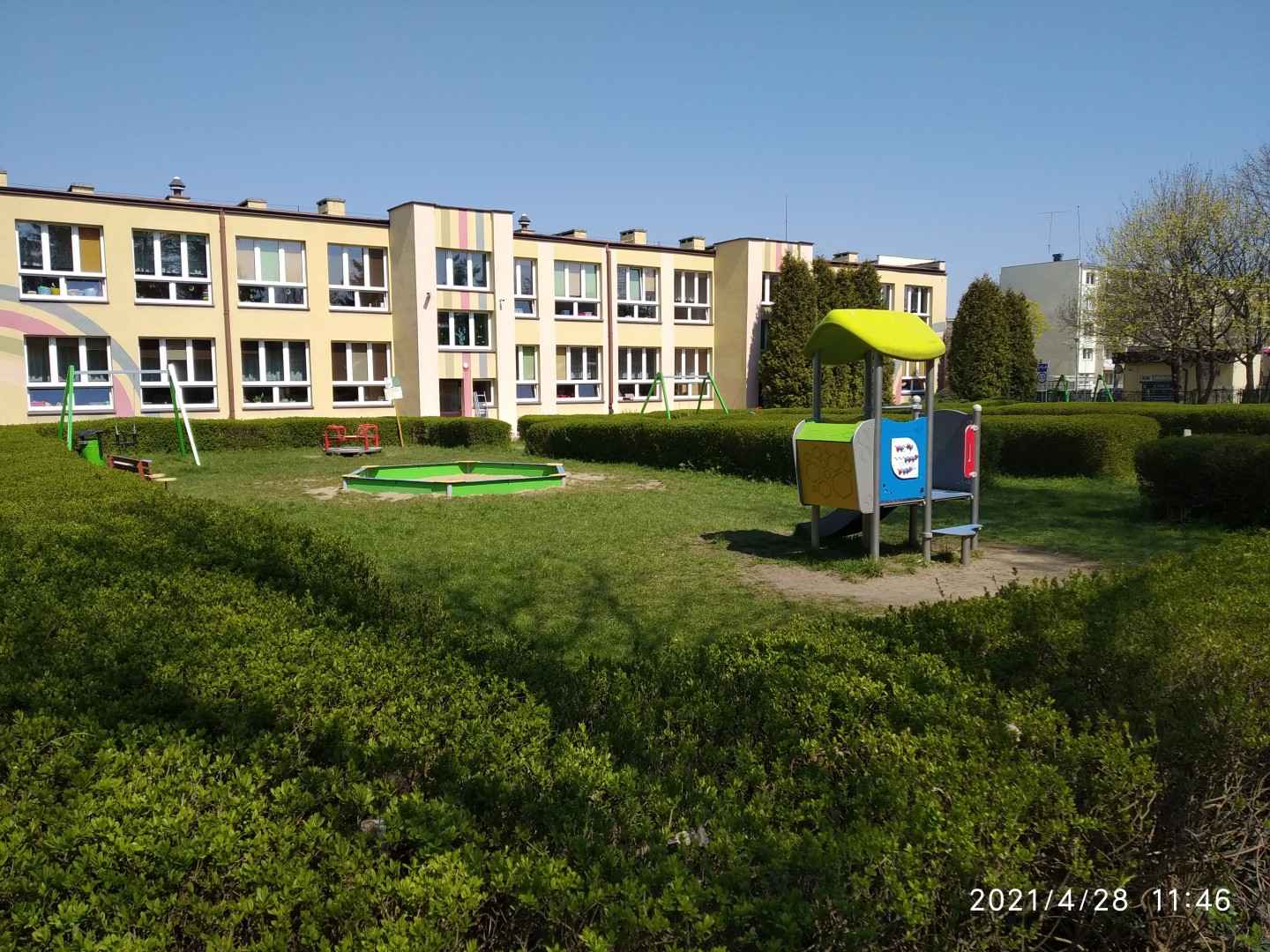Na zdjęciu widać zielony żywopłot, zabawki placu zabaw, a w oddali duży żółty budynek