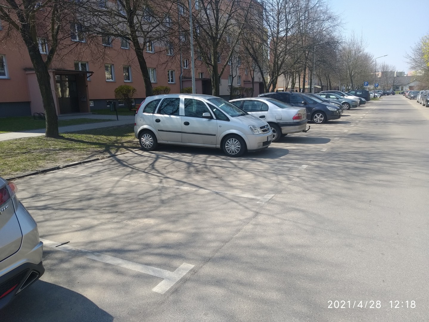 Na zdjęciu widać parking pod blokiem, samochody i drzewa