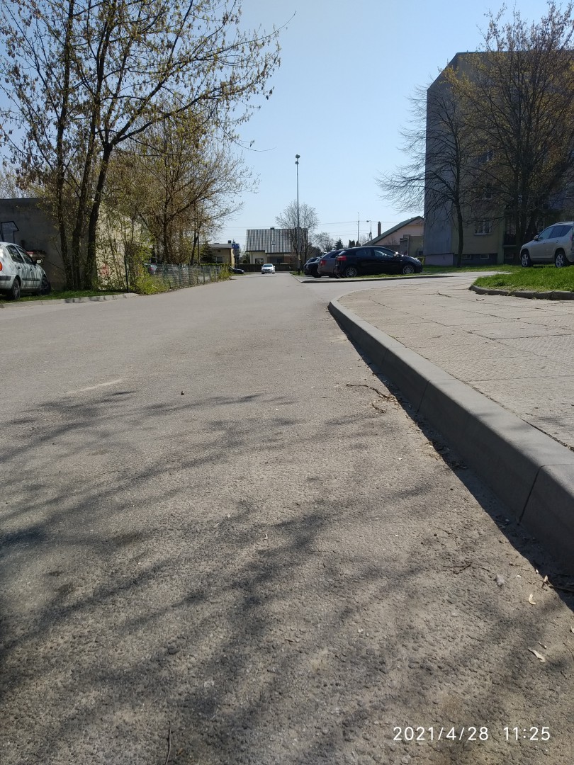 Na zdjęciu widać uliczkę, krawężnik, parkujące samochody, drzewa i bloki