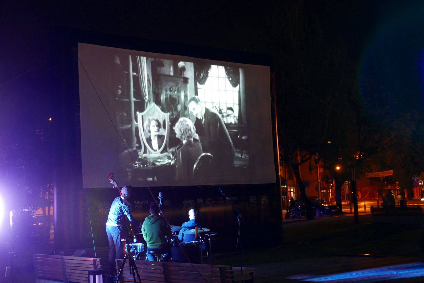 Zdjęcie przedstawia noc, duży ekran kinowy a na jego tle muzyków z instrumentami